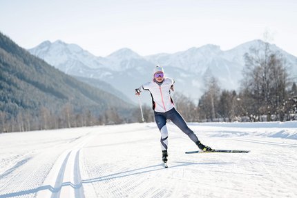 Olympische Winterspiele 2026 © Josef Plaickner Langläuferin in weißer Jacke auf verschneiter Loipe vor Bergen