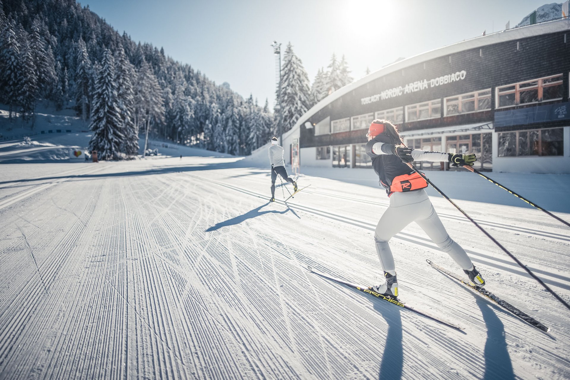 Cross Country Ski Holidays © Kottersteger Cross-country skiers skiing on a snowy trail near snowy forest and building