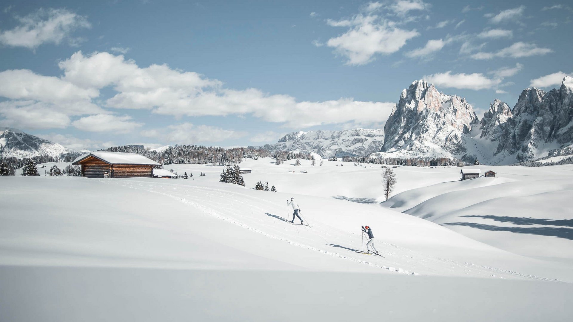 Langlauf Urlaub © Dolomiti Nordic Ski - Manuel Kottersteger Zwei Skifahrer in verschneiter Berglandschaft mit Hütten und Gipfeln