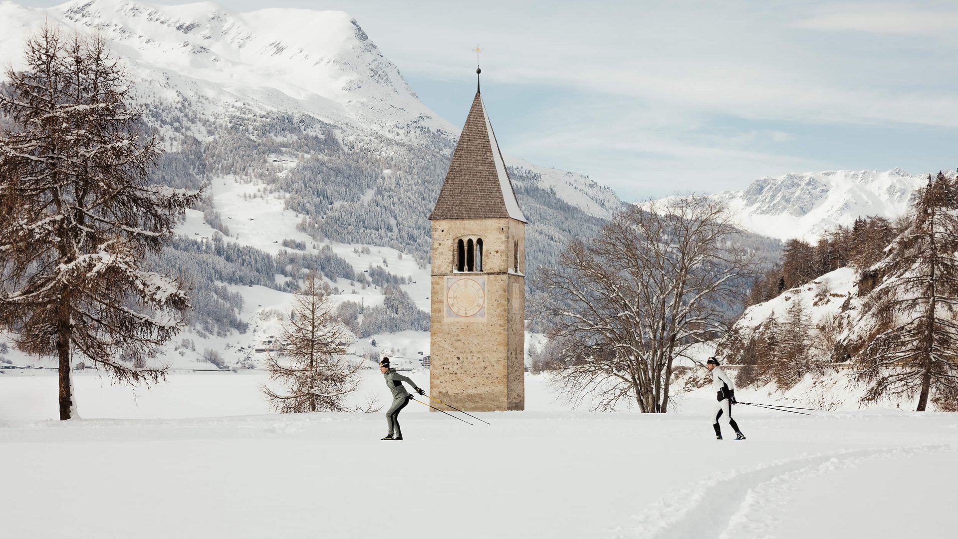 Langlauf Urlaub © Alex Moling Zwei Skilangläufer vor einem Kirchturm auf einer schneebedeckten Ebene mit Bergen