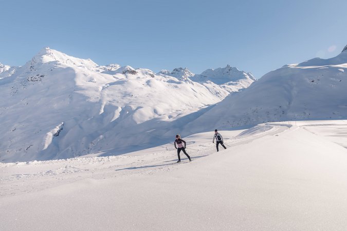 Langlauf Urlaub © TVB Paznaun-Ischgl Zwei Langläufer auf verschneiter Berglandschaft bei klarem Himmel