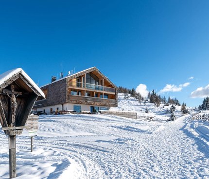 Alm-Hotel Starkenfeld Hütte © Jonas Eisenstecken Schneebedecktes Berghaus mit Winterweg und hölzernem Kreuz in den Alpen