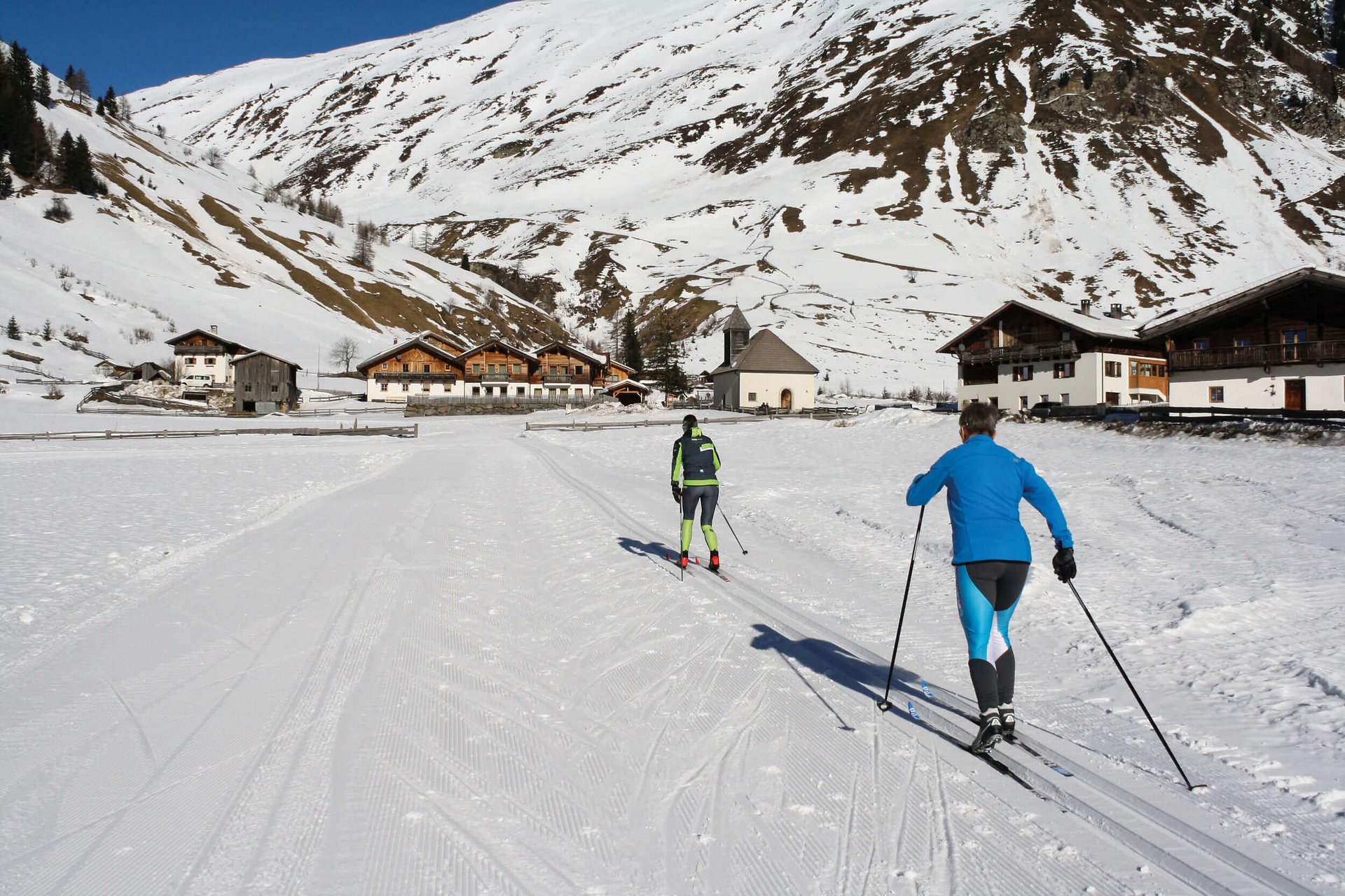 Cross Country Ski Holidays © Judith Vasselai Two cross-country skiers in snowy village with mountain backdrop