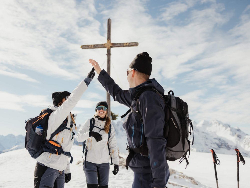 Langlauf Urlaub © Alex Moling Drei Wanderer geben sich im Schnee an einem Gipfelkreuz einen High Five