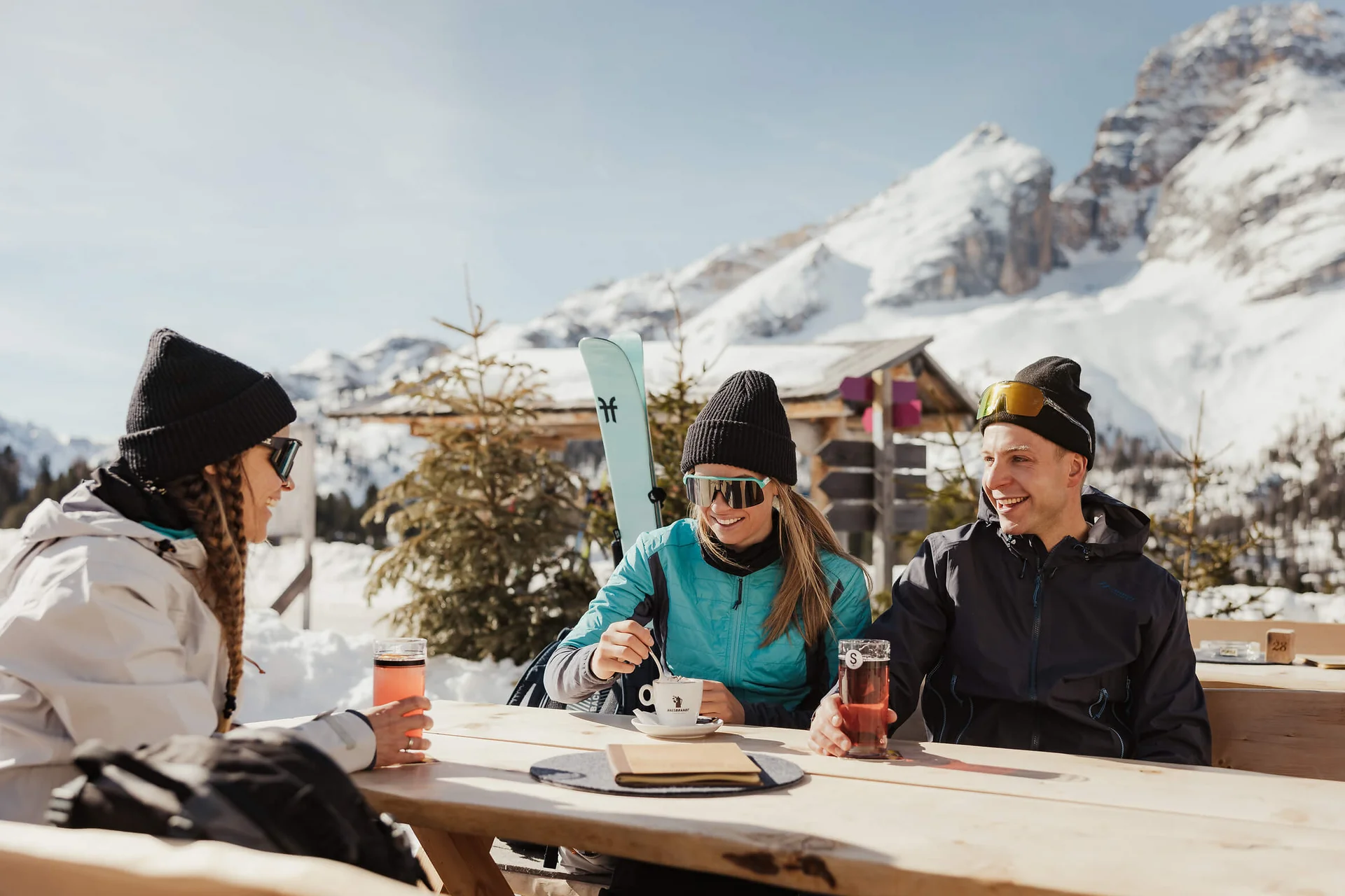 Langlauf Urlaub © Alex Moling Drei Personen genießen Getränke draußen mit verschneiten Bergen im Hintergrund