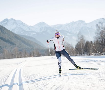 Olympische Winterspiele 2026 © Josef Plaickner Langläuferin in weißer Jacke auf verschneiter Loipe vor Bergen