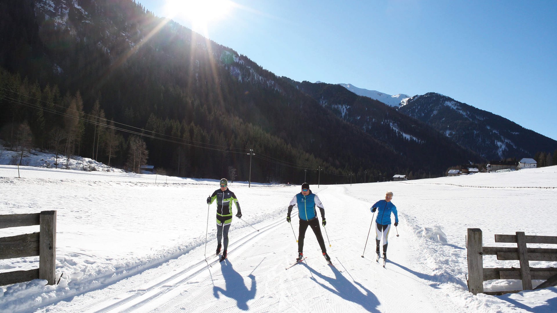 Langlauf Urlaub im Sarntal © Judith Vasselai Drei Langläufer auf schneebedeckter Strecke mit Bergen und Sonne im Hintergrund