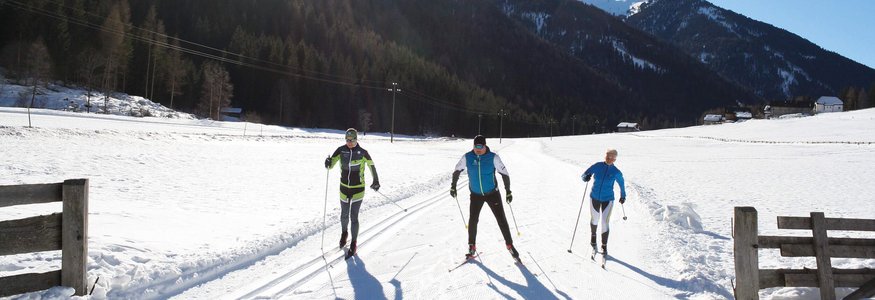 Sarntal © Judith Vasselai Drei Langläufer auf schneebedeckter Strecke mit Bergen und Sonne im Hintergrund