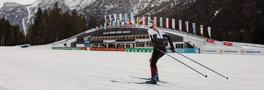 Apparthotel Germania *** © Alex Moling Skilangläufer vor verschneiten Bergen und Nordic Arena in Toblach
