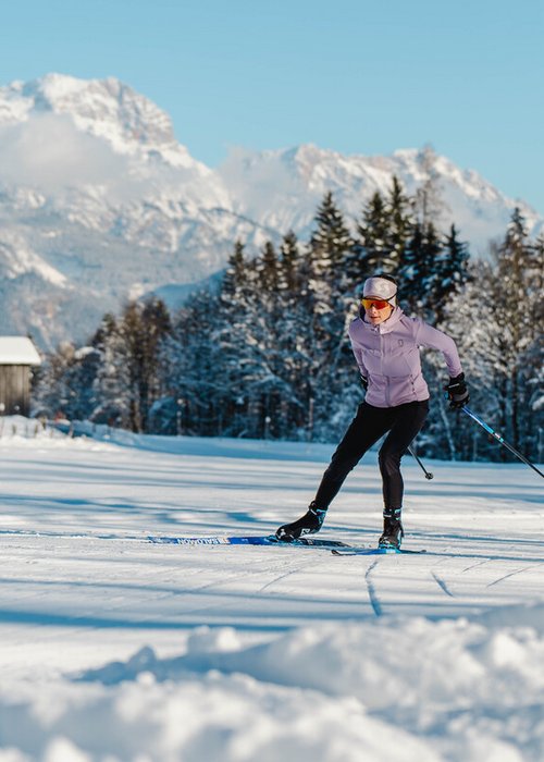 Langlauf Urlaub © Michael Geißler Langläuferin im Winter vor verschneitem Wald und Bergen