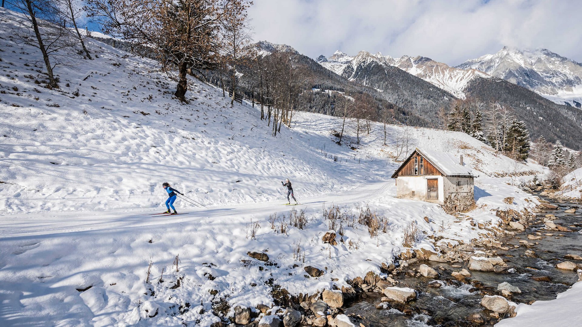 Langlauf Urlaub in der Dolomitenregion Kronplatz © Harald Wisthaler Zwei Skilangläufer auf verschneiter Bergstrecke neben Bach und Hütte