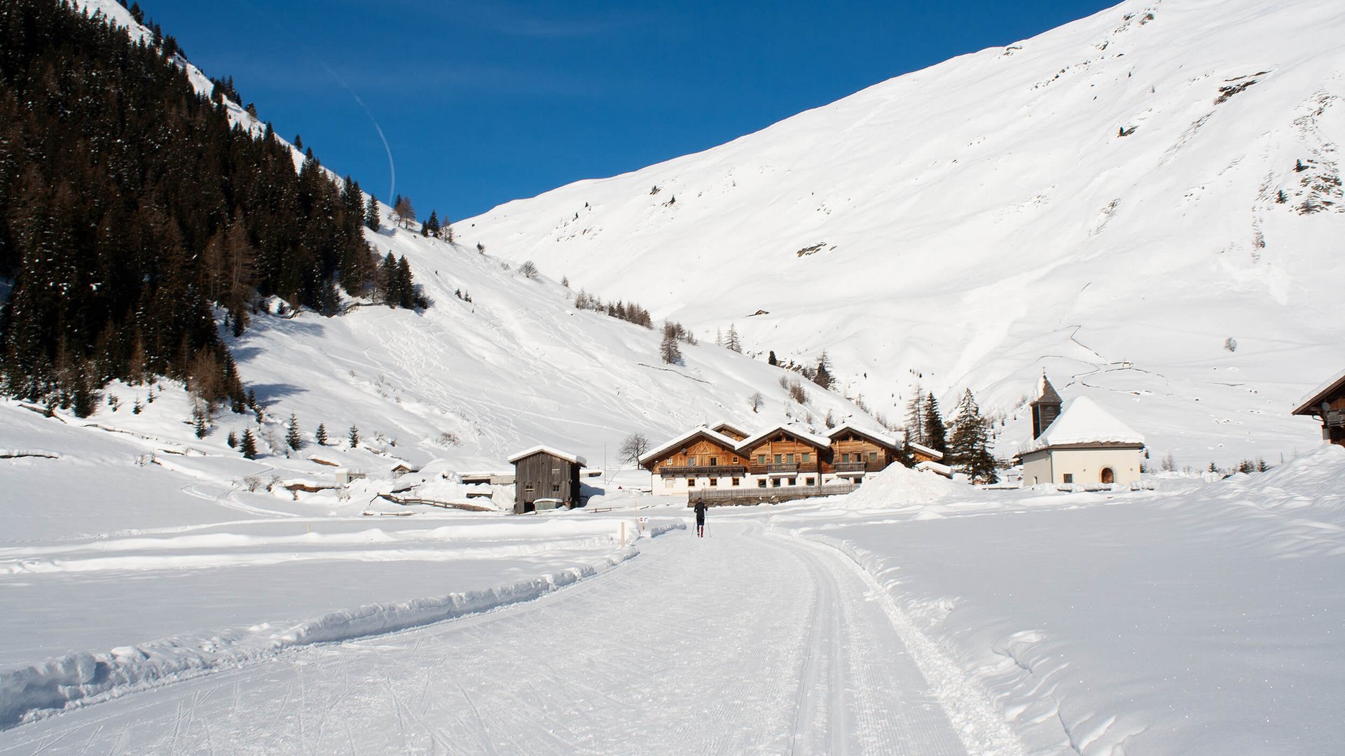 Langlauf Urlaub im Sarntal © Judith Vasselai Winterlandschaft mit Schlittenfahrt auf verschneiter Straße und Häusern vor Bergen