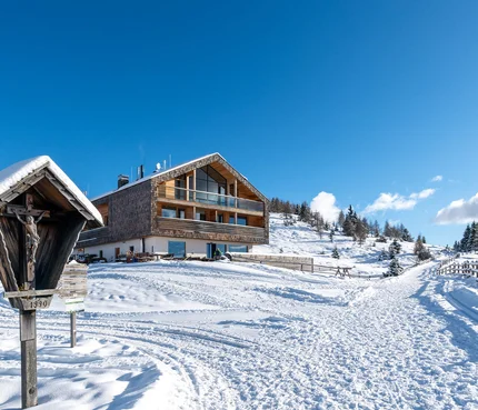 Alm-Hotel Starkenfeld Hütte © Jonas Eisenstecken Schneebedecktes Berghaus mit Winterweg und hölzernem Kreuz in den Alpen