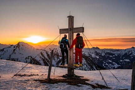 Ski touring days in the Bergsteigerdorf® Hüttschlag © TVB Großarltal / Peter Maier Two hikers standing at a summit cross during sunset in snowy mountains