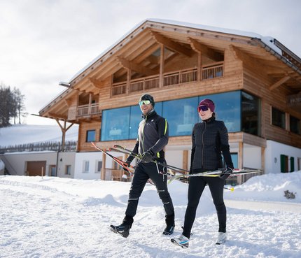 Hotel Ritsch © Fabian Dalpiaz Zwei Skifahrer tragen Langlaufskier vor einem Chalet im Schnee