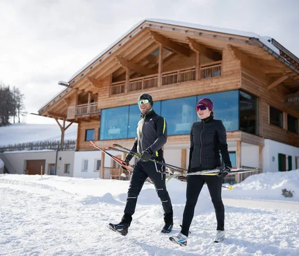 Hotel Ritsch © Fabian Dalpiaz Zwei Skifahrer tragen Langlaufskier vor einem Chalet im Schnee