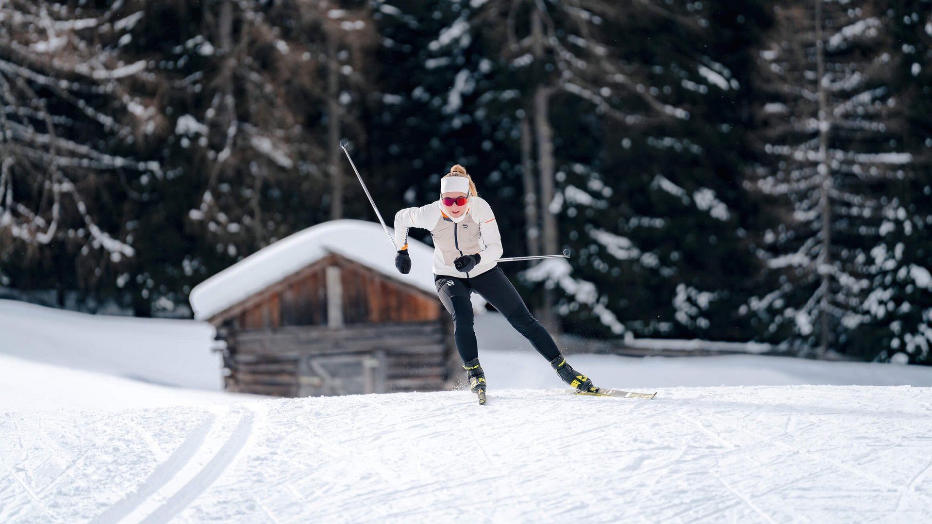 Langlauf Urlaub in der Dolomitenregion Kronplatz © Josef Plaickner Frau beim Langlaufen vor verschneiter Hütte und Wald