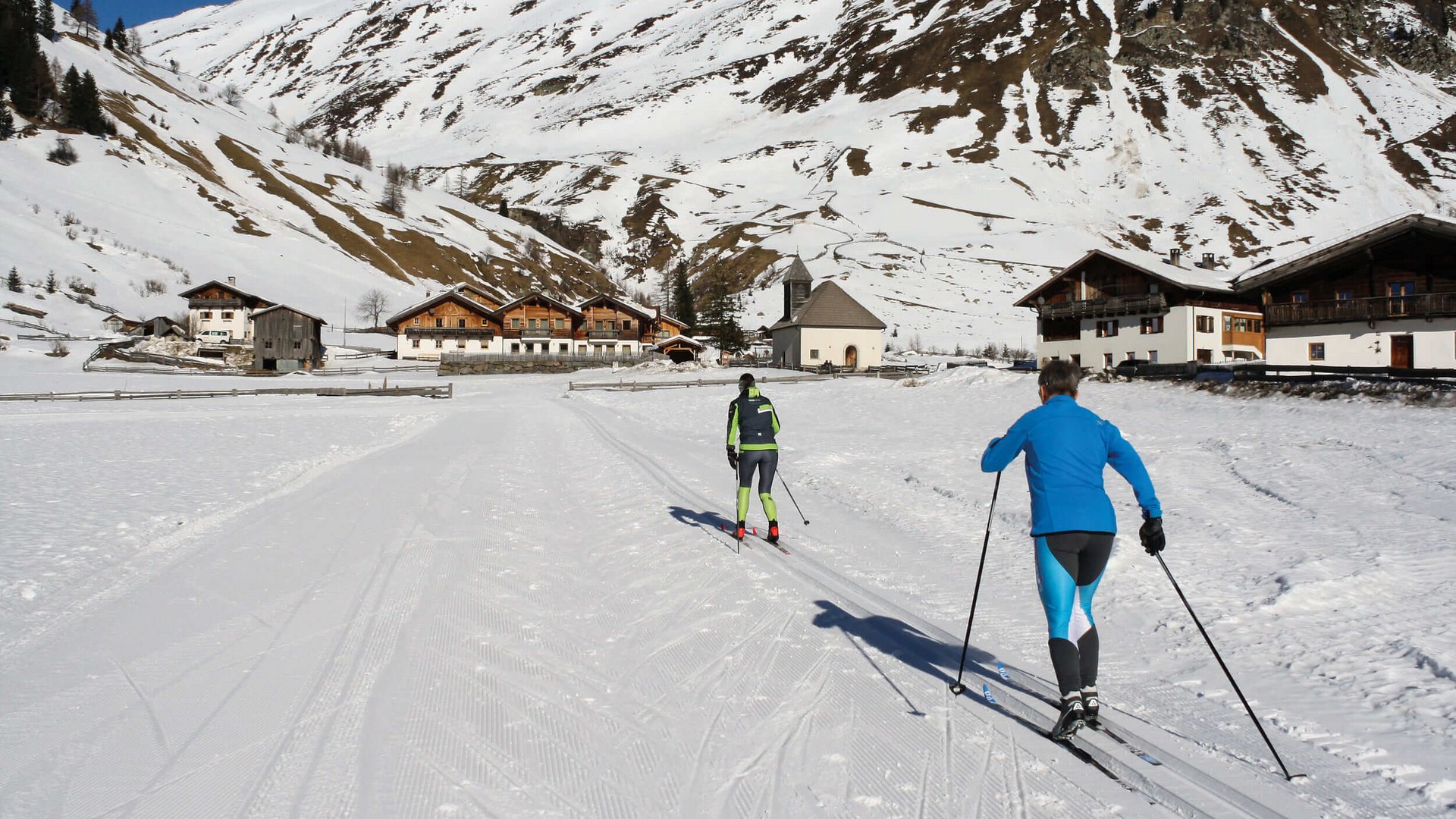 Langlauf Urlaub im Sarntal © Judith Vasselai Zwei Skilangläufer in verschneitem Dorf vor Bergkulisse