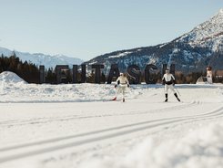 Region Seefeld - Tirols Hochplateau © Alex Moling Zwei Skifahrer beim Langlauf vor Leutasch-Schrift im Schnee mit Bergen im Hintergrund